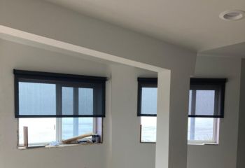 Interior view of a partially finished room featuring large windows with dark electric shades, a central support column, and unfinished flooring. The room is bright with natural light coming through the windows.